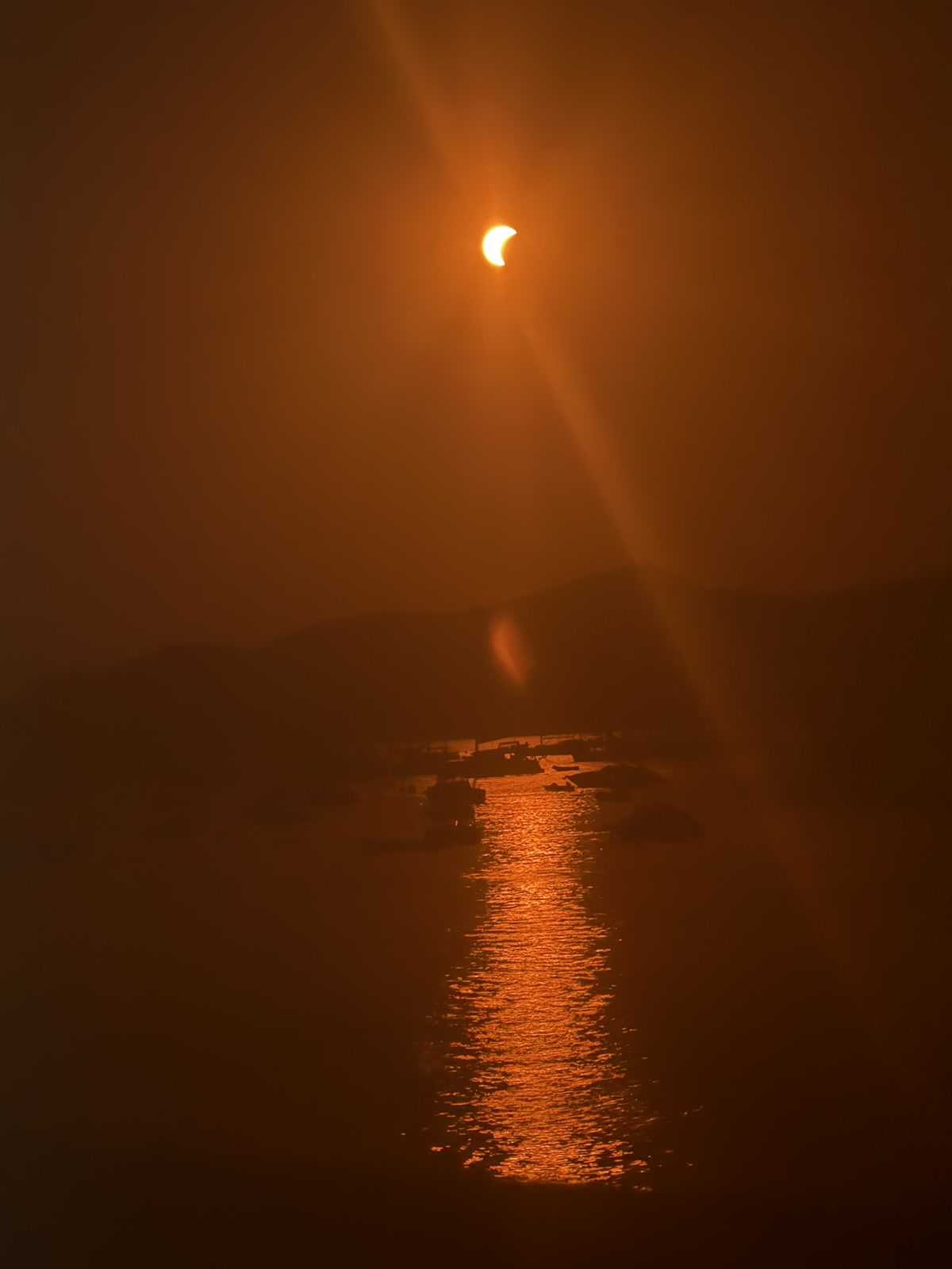 An orange crescent Sun above the sea with a long bright reflection reaching toward the shore.